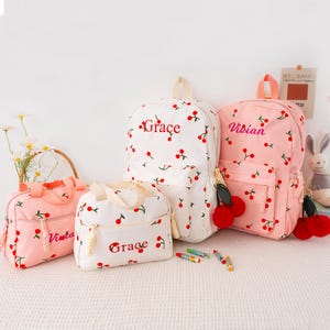 May include: A collection of cherry-printed backpacks and lunch bags in white and pink. The bags feature red cherry accents and the names "Grace" and "Vivian" embroidered in red. The bags have matching red pom-pom keychains.