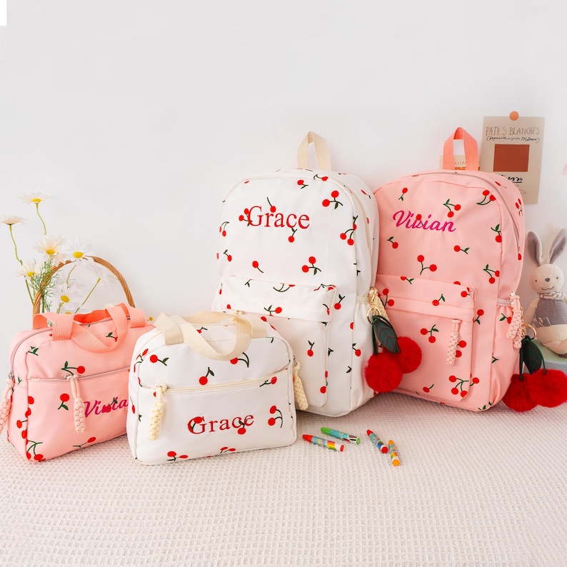 May include: A collection of cherry-printed backpacks and lunch bags in white and pink. The bags feature red cherry accents and the names "Grace" and "Vivian" embroidered in red. The bags have matching red pom-pom keychains.
