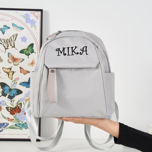 Puede incluir: Una mochila gris claro con el nombre "MIKA" bordado en negro en el bolsillo delantero. La mochila tiene una cremallera superior, un peque&ntilde;o bolsillo delantero y correas ajustables para los hombros. La mochila es sostenida por una persona.