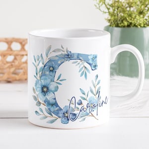 Può includere: Tazza in ceramica bianca con un motivo floreale blu e la lettera "C" con il nome "Caroline" scritto in corsivo.