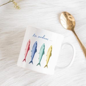 Può includere: Tazza in ceramica bianca con illustrazioni di quattro pesci colorati: rosso, blu, verde e giallo. La tazza presenta la scritta blu "Les sardines...". Un cucchiaio dorato è appoggiato accanto alla tazza.