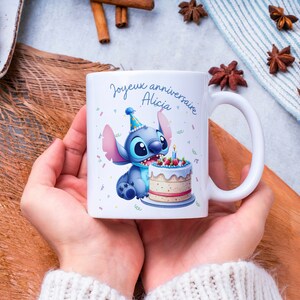 May include: White ceramic mug with a blue cartoon character holding a birthday cake. The mug has the text "Joyeux anniversaire Alicia" on it.