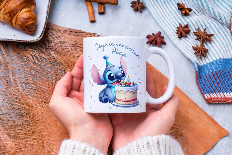 May include: White ceramic mug with a blue cartoon character holding a birthday cake. The mug has the text "Joyeux anniversaire Alicia" on it.