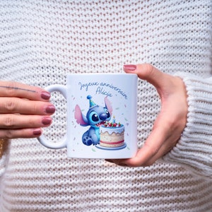 May include: White ceramic mug with a blue cartoon character holding a birthday cake. The text on the mug reads "Joyeux anniversaire Alicia".