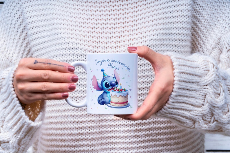 May include: White ceramic mug with a blue cartoon character holding a birthday cake. The text on the mug reads "Joyeux anniversaire Alicia".