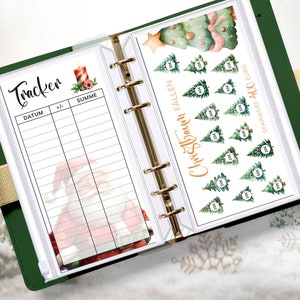 May include: A white planner with a green cover and gold rings. The planner has a page with a Christmas tree savings tracker and a page with a Christmas tracker for recording dates and amounts. The Christmas tree savings tracker has a watercolor illustration of a Christmas tree with ornaments and a star on top. The Christmas tracker page has a watercolor illustration of Santa Claus.