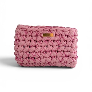 May include: A rectangular crochet clutch bag made from thick, pink fabric yarn. The bag features a chunky, textured knit pattern and a small, rectangular gold-toned metal plaque centered on the front with the text LULU engraved on it.