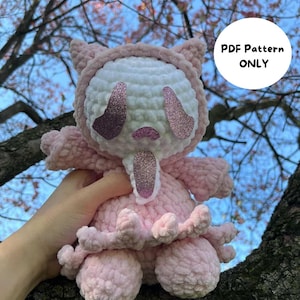 May include: A handmade pink and white crochet plush toy with glitter accents. The toy has a hood with ears, large glittery eyes, and a small mouth. The text "PDF Pattern ONLY" is visible in a white circle. The toy is set against a background of tree branches and a blue sky.