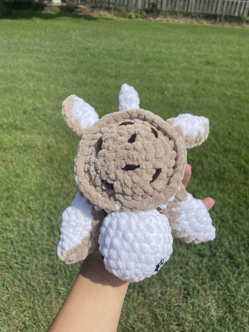 May include: A crocheted plush turtle toy with a brown shell and white legs and head. The turtle has a smiling face.