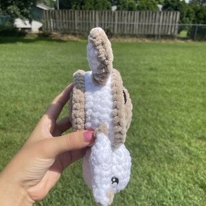 May include: A crocheted stuffed animal in the shape of a bunny. The bunny is white with brown accents and has a black button eye. The bunny is being held by a hand.