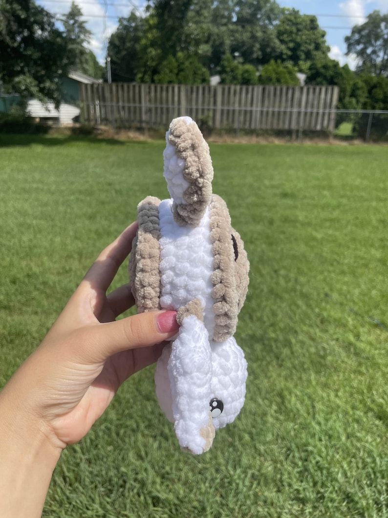 May include: A crocheted stuffed animal in the shape of a bunny. The bunny is white with brown accents and has a black button eye. The bunny is being held by a hand.