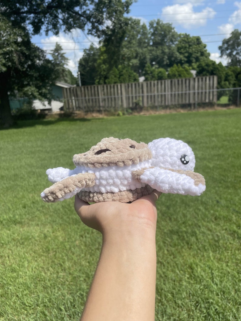 May include: A crocheted stuffed turtle toy with a brown shell and white head and legs. The turtle is being held in a person's hand.