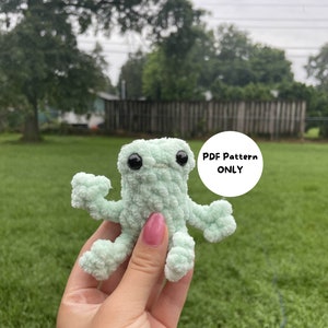 May include: A crocheted green frog amigurumi toy with black eyes. The toy is being held in a hand. The text "PDF Pattern ONLY" is visible in the image.