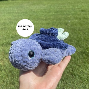 May include: A crocheted blue and green turtle plush toy. The toy has a large, round head with a single black eye. The toy is being held in a person's hand. The text "PDF PATTERN ONLY" is visible in a white circle.