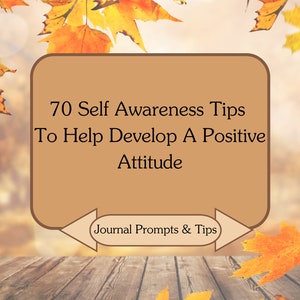 May include: A brown rectangle with the text "70 Self Awareness Tips To Help Develop A Positive Attitude" and "Journal Prompts & Tips". The rectangle is surrounded by a blurred background of autumn leaves and a wooden surface.