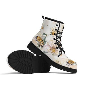 May include: White combat boots with a floral and bee design. The boots feature a black sole, black laces, and a pattern of white flowers and honeybees. The boots are designed for all-weather wear.