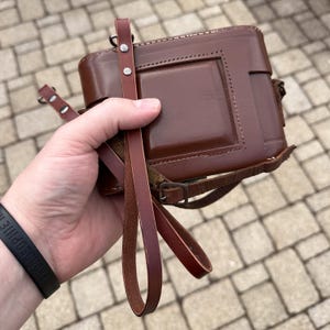 Könnte beinhalten: Eine braune Leder-Kameratasche mit passendem Riemen. Die Tasche hat ein rechteckiges Design mit einer genähten Bordüre und einer quadratischen Vertiefung auf der Vorderseite. Der Riemen ist mit Metallnieten und einer Schnalle befestigt.
