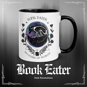 May include: White ceramic mug with a black interior and handle. The mug features a circular design with a skeletal creature curled around a book, surrounded by floral elements. Text reads "Book Eater" and "Devourer of Worlds".