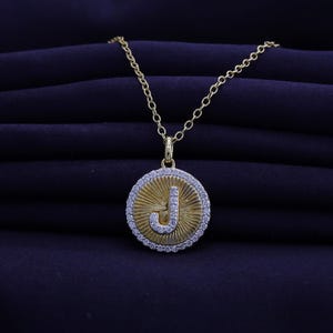 May include: A gold-colored pendant necklace featuring the letter "J" in a circular design. The pendant has a sunburst pattern and is encircled by a row of small, clear stones. The necklace chain is also gold-colored.