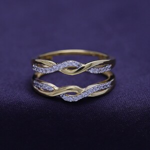 Op de afbeelding: Een gouden ring met een gedraaid ontwerp en kleine, heldere stenen. De ring is ontworpen om te worden gedragen als een ringversterker.