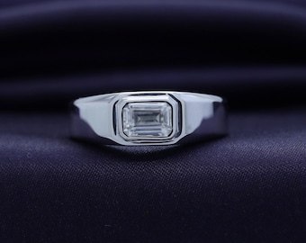 May include: A silver ring with a rectangular, emerald-cut gemstone. The ring has a polished finish and a simple, modern design. The gemstone is clear and reflects light.