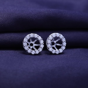 May include: A pair of silver-coloured earring jackets, each featuring a halo of small, clear gemstones. The jackets have a starburst design in the centre. They are presented on a dark blue fabric background.