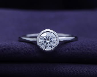 May include: A silver ring with a large, round, clear gemstone set in a bezel setting. The ring is displayed on a dark purple fabric, highlighting the sparkle of the stone. The band is thin and polished.