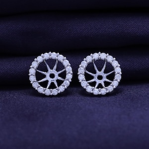 May include: A pair of silver-toned earring jackets. Each jacket is circular, with a central starburst design and a surrounding halo of small, clear gemstones. The earrings are displayed against a dark blue background.
