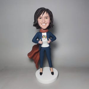 Könnte beinhalten: Eine personalisierte Bobblehead-Figur einer Frau mit kurzen schwarzen Haaren, die eine blaue Anzugsjacke, ein weißes Hemd mit einem bunten Logo, einen roten Schal und einen roten Umhang trägt. Die Figur steht auf einem weißen Sockel.