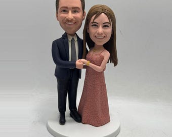 May include: A wedding cake topper featuring a smiling couple. The man wears a navy suit with a gray tie, and the woman is in a pink dress. They are holding hands, standing on a white base.