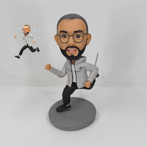 Può includere: Una statuetta di un uomo che corre con occhiali, barba e felpa grigia. La figura è su una base circolare grigia. Una versione più piccola della statuetta è sullo sfondo, anch'essa in corsa. La statuetta indossa pantaloni e scarpe neri.