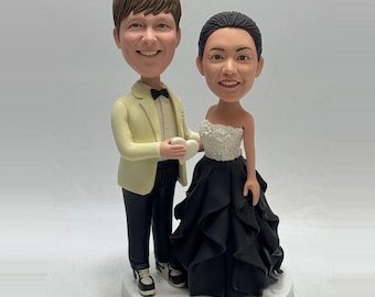 May include: A wedding cake topper featuring a smiling couple. The man wears a light yellow suit jacket, black pants, and sneakers. The woman wears a strapless black gown with a white lace bodice. They are holding a white heart.