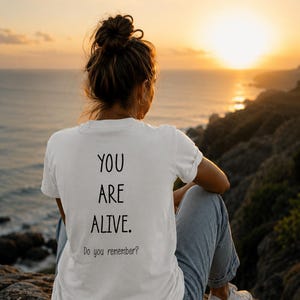 Könnte beinhalten: Ein weißes T-Shirt mit den schwarzen Aufdruck "YOU ARE ALIVE. Do you remember?". Das T-Shirt wird von einer Person getragen, die auf einem Felsen sitzt und einen Sonnenuntergang über dem Meer betrachtet. Die Person trägt eine blaue Jeans und weiße Turnschuhe.