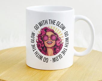 Może przedstawiać: Biały ceramiczny kubek z grafiką kobiety z różowymi włosami i okularami przeciwsłonecznymi, otoczony tekstem "GO WITH THE GLOW". Kubek stoi na drewnianym stojaku na białym, puszystym tle.