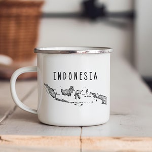 May include: White enamel mug with a silver rim and handle. The mug features the word "INDONESIA" in black, above a detailed black and white map outline of the country. The mug is on a light-colored wooden surface.