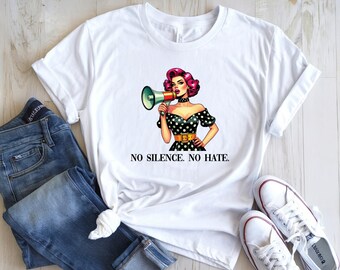 May include: White t-shirt featuring a pop art style illustration of a woman with pink hair holding a megaphone. She wears a black polka dot dress and a choker. The text "NO SILENCE. NO HATE." is printed below the image. Blue jeans and white sneakers are also in the image.
