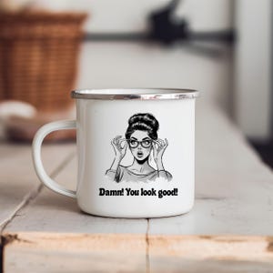 Könnte beinhalten: Weißer Emaillebecher mit silbernem Rand und Henkel. Der Becher zeigt eine Schwarz-Weiß-Illustration einer Frau mit Brille, darunter der Text "Damn! You look good!" gedruckt.
