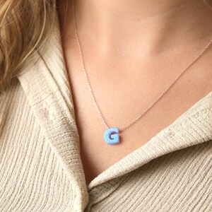May include: A silver necklace with a light blue letter "G" pendant. The pendant is a solid color and hangs from a delicate chain. The necklace is worn against a beige collared shirt.