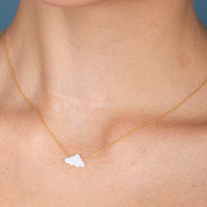Puede incluir: Un delicado collar dorado con un pequeño colgante en forma de nube blanca. La nube tiene una apariencia texturizada, similar al ópalo. El collar se muestra en el cuello de una persona, mostrando el diseño y el estilo de la joya.