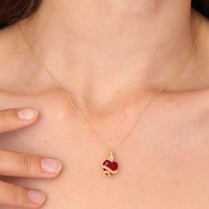 Peut inclure: Un collier en or avec un pendentif en forme de cœur orné d'une pierre rouge. Un motif de serpent doré entoure le cœur. Le collier est porté autour du cou d'une personne.