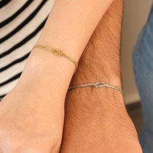 K&ouml;nnte beinhalten: Zwei passende Armb&auml;nder, eines goldfarben und eines silberfarben, jeweils mit einem Knotendesign. Die Armb&auml;nder werden an Handgelenken getragen, ein Arm in einem gestreiften Oberteil und der andere in einem Jeanshemd.