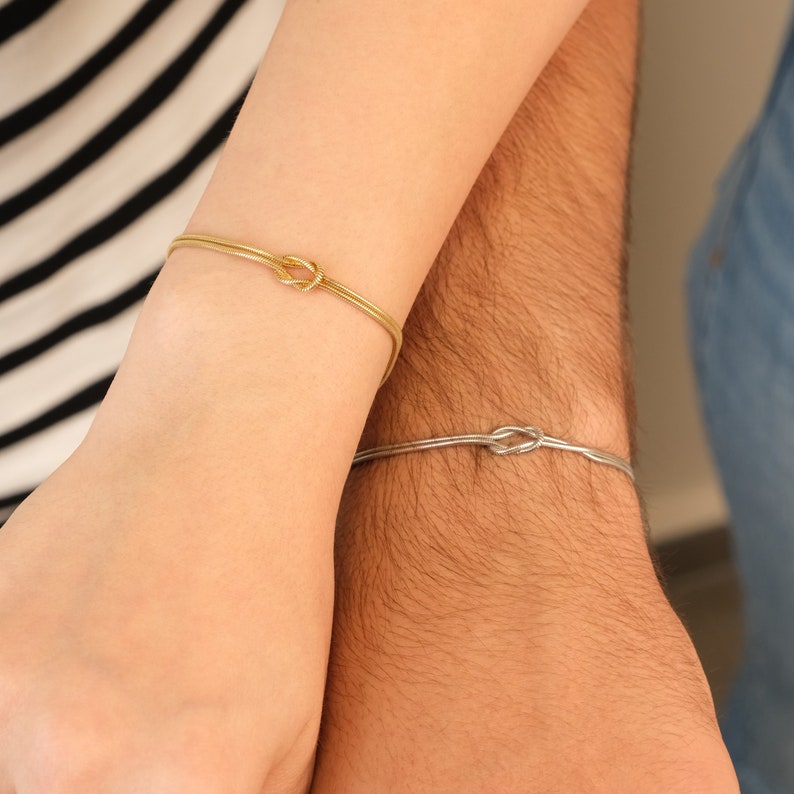 K&ouml;nnte beinhalten: Zwei passende Armb&auml;nder, eines goldfarben und eines silberfarben, jeweils mit einem Knotendesign. Die Armb&auml;nder werden an Handgelenken getragen, ein Arm in einem gestreiften Oberteil und der andere in einem Jeanshemd.
