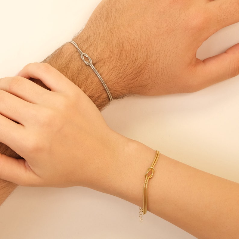 K&ouml;nnte beinhalten: Zwei Armb&auml;nder, eines silberfarben und eines goldfarben, jeweils mit einem Knoten-Design. Die Armb&auml;nder werden an Handgelenken getragen und zeigen ihren minimalistischen Stil. Das goldene Armband hat eine Kettenverl&auml;ngerung.