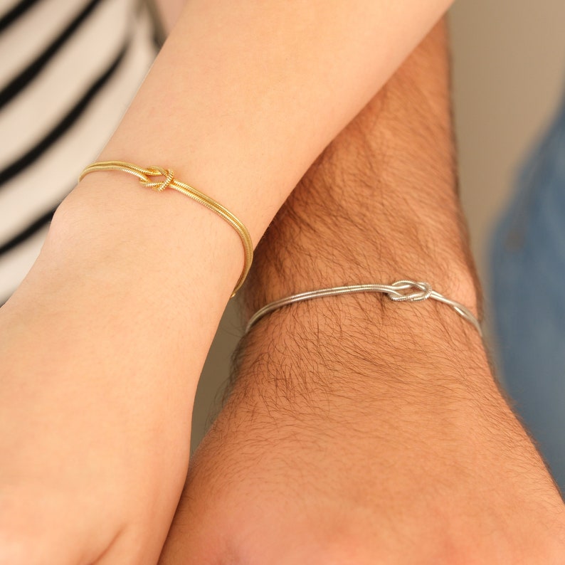 K&ouml;nnte beinhalten: Zwei passende Armb&auml;nder, eines goldfarben und eines silberfarben, jeweils mit einem Knotendesign. Die Armb&auml;nder werden an Handgelenken getragen, wobei ein Arm im Vordergrund und der andere im Hintergrund ist. Das goldfarbene Armband ist links, das silberfarbene rechts.