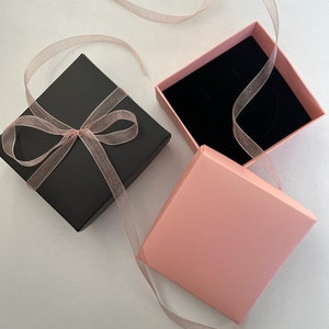 K&ouml;nnte beinhalten: Eine schwarze quadratische Geschenkbox, gebunden mit einem durchsichtigen rosa Band, neben einer offenen rosa Box mit schwarzem Samtinnenraum. Ein rosa Band ist &uuml;ber die Boxen drapiert, was auf eine Pr&auml;sentation f&uuml;r Schmuck oder ein kleines Geschenk hindeutet.