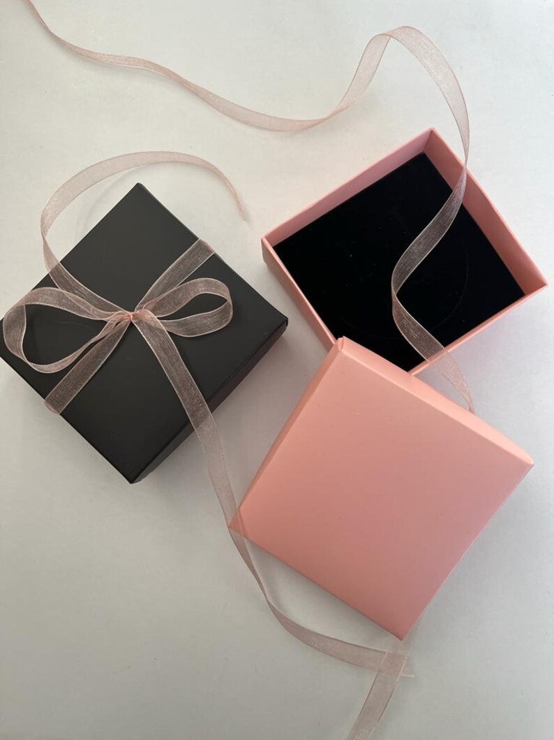 K&ouml;nnte beinhalten: Eine schwarze quadratische Geschenkbox, gebunden mit einem durchsichtigen rosa Band, neben einer offenen rosa Box mit schwarzem Samtinnenraum. Ein rosa Band ist &uuml;ber die Boxen drapiert, was auf eine Pr&auml;sentation f&uuml;r Schmuck oder ein kleines Geschenk hindeutet.