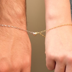 Könnte beinhalten: Zwei passende Armbänder mit einem herzförmigen Anhänger. Eines ist silberfarben, das andere goldfarben, und der Herzanhänger ist zwischen beiden geteilt. Die Armbänder sind durch eine Kette verbunden und werden an den Handgelenken getragen.