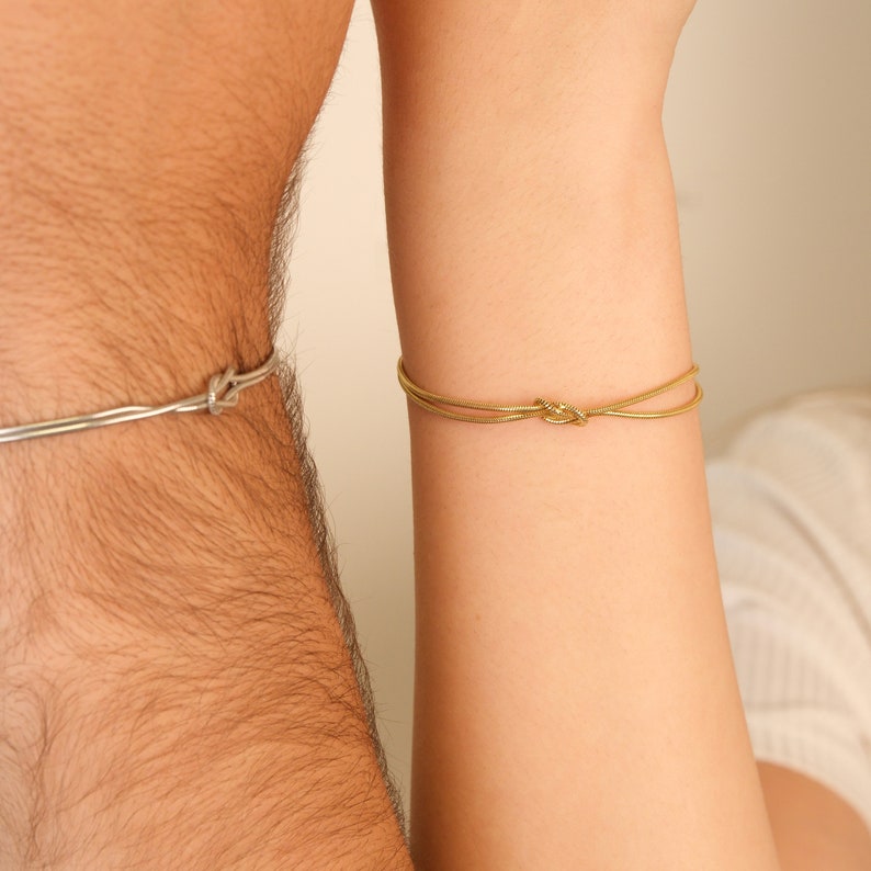 K&ouml;nnte beinhalten: Zwei Armb&auml;nder, eines silberfarben und eines goldfarben, jeweils mit einem Knoten-Design. Das silberne Armband befindet sich an der Taille einer Person, w&auml;hrend das goldene Armband am Handgelenk ist. Die Armb&auml;nder sind schlicht und elegant.