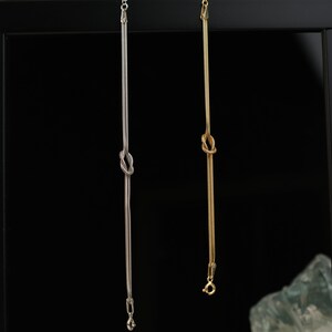 K&ouml;nnte beinhalten: Zwei filigrane Halsketten, eine silberfarbene und eine goldfarbene, h&auml;ngen vor einem schwarzen Hintergrund. Jede Kette hat einen Knoten in der Mitte und eine feine Kette. Die silberne Kette hat einen kleinen Anh&auml;nger, die goldene einen Verschluss.