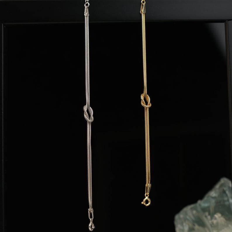 K&ouml;nnte beinhalten: Zwei filigrane Halsketten, eine silberfarbene und eine goldfarbene, h&auml;ngen vor einem schwarzen Hintergrund. Jede Kette hat einen Knoten in der Mitte und eine feine Kette. Die silberne Kette hat einen kleinen Anh&auml;nger, die goldene einen Verschluss.
